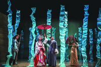 A color photo of a scene on stage. A male figure is holding an umbrella on the right of a female figure. Two other figures are present, each standing on one side of the two central figures. More than a dozen long blue drapes are hanging vertically from the ceiling, symbolizing rain.