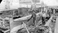 A black and white photograph of two men and one barefoot child standing beside a long dugout canoe. The unifinished canoe is held up by a wooden structure.