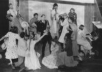 Vilna Ghetto residents dance in a circle around SS commandant Kittel, who plays the saxophone. Several other musicians stand or sit behind the dancers in the background, playing the bass, an accordion, clarinet, violin, and piano.