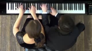 Two players sit at a grand piano and play together animatedly.