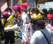As part of a protest march, a black woman stands blindfolded, carrying a sword and scales, her white gown bloody at the hem. She is surrounded by protestors carrying flags, bullhorns, and handmade signs reading, “Equitable Redistribution of Land” and “We demand a society that’s free from rape.”