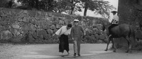 In this black and white capturing of a rural town, the teen protagonist and his school friend stroll away, in center frame, from a very clean and regular stone wall (evidently a temple grounds), which stretches from Left to Right, dominating the background, whilst a peasant on a horse ambles away, screen right, past the sharp corner of an even higher wall across the street (on Screen Left).