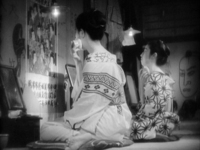Two women kneel while applying makeup, facing a wall covered in calligraphy and torn posters.