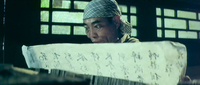 Image of a man standing behind a wide piece of paper that has black calligraphy painted on it. The paper is so thin that the viewer can see the words through the other side.