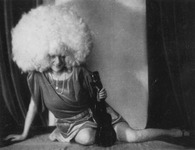 Black-and-white photograph of performer Zerline Balten, a European woman wearing her hair as a large blond Afro and holding a ukulele. She smiles, sits with a leg stretched out behind her in front of curtains, and wears a short shiny rayon dress and beaded necklace.