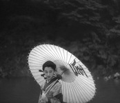 A woman in a kimono with a paper parasol, with calligraphic text written on it, standing in front of trees.