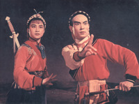 Close-up shot of a man and woman in red costumes looking into the distance. The man points forward with his index and middle fingers raised. The woman has a sword slung over her shoulder.
