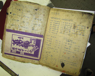 A photo of an old and yellowed book opened to pages showing charts and details of calligraphic characters. A purple and white pamphlet has been paper clipped to the interior of the left page.