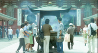 People gather around a large incense burner in a courtyard, framed in the background by a temple gate and large calligraphic banners.