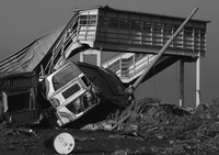 An overturned train car has knocked aside a telephone pole, amidst debris and rubble. A second train car is overturned to the left of it. Further back, a covered elevated walkway and stairs can be seen, partially collapsed.