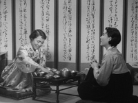 Two women sit at a chabudai in front of room dividing screen with black calligaphy printed on it, in black and white cinematography.