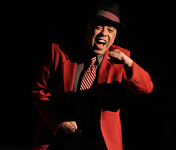 Merced smiles broadly, wearing a black hat with polka-dot trim, a suit, and a striped tie. Has fist in mid-air, appears to be dancing.