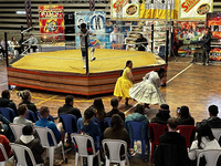 A referee stands inside a six-­sided wrestling ring with banners for the promotion behind. Two women grapple outside the ring—­one wearing a yellow blouse and pollera skirt, and the other a white blouse and pollera skirt. Audience members on white plastic chairs look on.