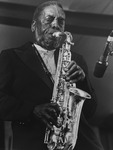 Blackandwhite image depicting a man standing on a stage, playing an alto saxophone intensely with his eyes closed, its bell to the viewer’s right.