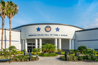 Photograph of the exterior front entrance of the American Police Hall of Fame and Museum, a large white building with a blue sign above the front door and four white pillars on each side of the front door. There are several palm trees, two benches, a paved path, and landscaped plants in front of the museum.