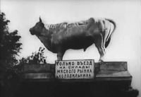 Large statue of a bull on pedestal with inscription in Russian (no translation provided in film), with trees in the background to the left.