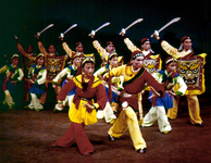 Three rows of dancers in bright yellow, red, and blue costumes line the stage. In the back, men hold large sabers above their heads and shields painted with lion faces. In the front, the dancers stand cross-legged and hunch forward as if advancing.