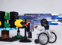 Color photo: a Lego person in a white long-­sleeve shirt, wheelchair, and long, dark hair, holds their left hand in front of the AUMI screen on an iPad on a stand.