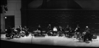 Black and white photo, thirteen people sit in a semi-­circle on a stage. Many performers are using AUMI, other instruments are present.