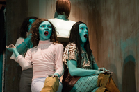 Two actors, wearing fashionable female clothes, long-­hair wigs, and makeup, covered in aqua-­blue body paint, sit together, mouths agape with a look of utter surprise.