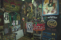 A range of different religious trinkets and posters in a stall in Takiye, where Shias mourn Hussain (the grandson of Muhammad the prophet) and Iranian martyrs. At least fifteen posters made from paper, cloth, and other materials cover the two walls visible, running from the floor to the roof in a disordered fashion. Various trinkets are hung on the walls, including beads, large white feathers, and fake flowers. A red, gold, and white patterned rug covers the floor, on which stand two small shoe racks containing dust-­covered black shoes.