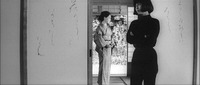 A film still showing an open sliding door, with calligraphic text written on both sides of the door and doorway. A woman in black clothes stands in the doorway, with a woman in a kimono standing further beyond.