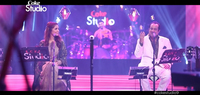 Long shot of singers Momina Mustehsan and Rahat Fateh Ali Khan in the Coke Studio version of “Afreen Afreen.” Seated to the right, Khan sports a white suit and sings with his eyes closed; Mustehsan looks on from the left, smiling. Mics, music stands, and sound monitors occupy the foreground; the drummer sits in the background, between the singers.