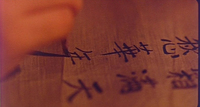 Close-up image of someone writing black calligraphy in pencil on a piece of wood.