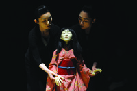 A puppet in a kimono is centred between two actresses in black, each holding one of the puppet’s wrists and supporting its body from the back, against a black background.