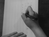 An individual writes black calligraphy on ruled paper, in black and white cinematography.