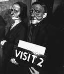 A woman and man in dark clothing lean against a wall. They wear half-­face masks with furrowed brows. The man holds a board bearing the legend “Visit 2.”