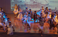 Color photograph of an ensemble of dancers from Fiji in formation on an indoor stage. Behind them is a projected image of a voyaging canoe.