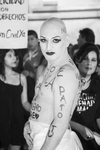 A seminaked person covered in white paint and heavy face makeup looks at the camera. On the person’s body are inscriptions of homophobic slurs.