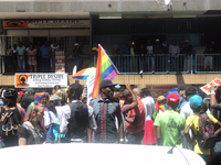 A group of around two dozen people march in the street, a rainbow flag prominently waving. A row of spectators look down from the balcony above.
