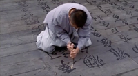 A man kneels on floorboards covered with calligraphic text, carving at the characters.