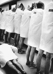 Eight men in knee-­length white smocks and flip-­flops are lined up with their backs to the camera, their palms pressed high against the wall they are facing. A ninth man is visible in the foreground, executing push-­ups.