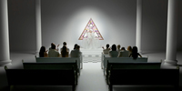 Poppy stands on an altar in white tulle prayer robe in front of a pink stained-­glass P. The first two rows of pews are filled.