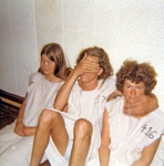 Three young women sit on the floor, their backs leaning against a blank, white wall. They are dressed uniformly in white smocks. The number 416 is visible at the shoulder of the woman on the right.
