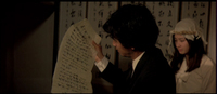 Man reads from a sheet of scroll paper, a calligraphic screen in the background.