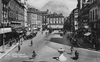 A postcard showing a photograph of a street view of Place Grenette, Grenoble. The street is mostly empty except for a trolley and several bicyclists. The street is lined with buildings four to five stories tall.