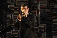 Solo performer with a Cesar haircut, wearing a black hoodie and holding a microphone close to his face. He stands against a wall of books from the bookstore he works at in the play.