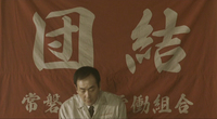 A man at the bottom of the frame speaks, with a large red banner with white callipgraphic text behind him
