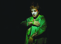A color photo of a stage performance. It features a figure in a green robe against a dark background. The figure wears bright pinkish red face paint and dazzling gold jewelry decorated with colorful stones.