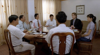 A framed calligraphic poem adorns the wall behind the white protagonist, who sits at a round dinner table with six Chinese people.