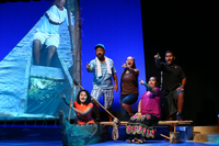 Color photograph of five performers on a theatrical set-piece of a Micronesian outrigger canoe. They are pointing and speaking in unison.
