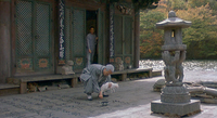 A squatting man paints calligraphy on floorboards with a white cat's tail, in front of a building, as another person watches.