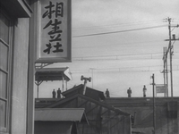 Calligraphy on a sign in the foreground, with buildings, power lines, and people waiting for a train in the background.