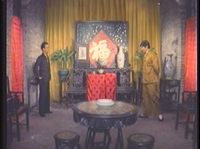 Two men face each other in a room with chairs and table, with a large calligraphic character on the wall behind them.