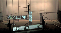 A dancer with arms clasped overhead balances on a metal structure that looks like scaffolding. Three panels with black-­and-­white images of faces extend on parts of the structure.