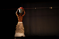 Fig. 2. Female dancer, muscular brown bare back to camera, long tulle and burlap skirt pinned with clothespins. Her arms are raised, holding a steel bucket of red laundry on her head.